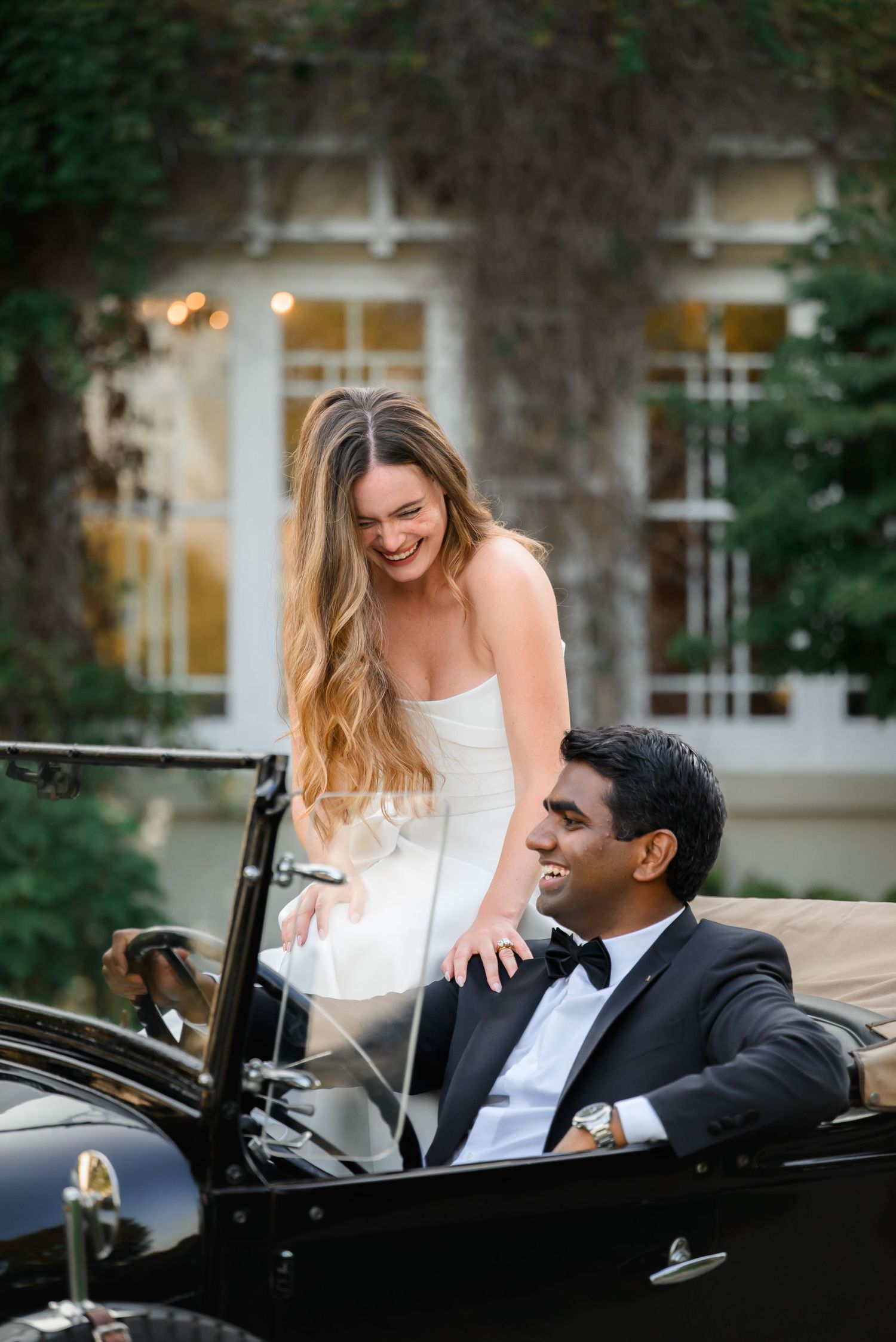 Bride in white gown laughs with groom in tuxedo seated in vintage black convertible car outside elegant venue.