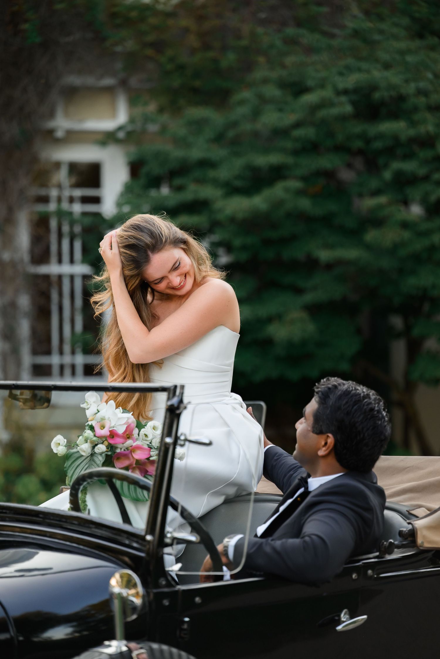 Bride in white dress laughs joyfully with groom in black suit seated in vintage convertible car.