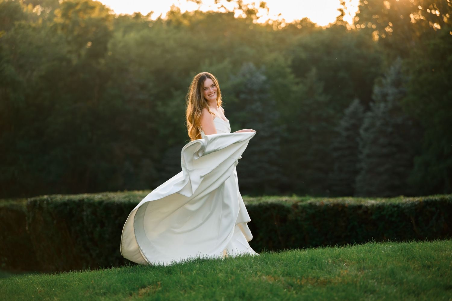 Bride in flowing white gown twirling on green lawn at golden hour with lush hedges and trees backdrop.