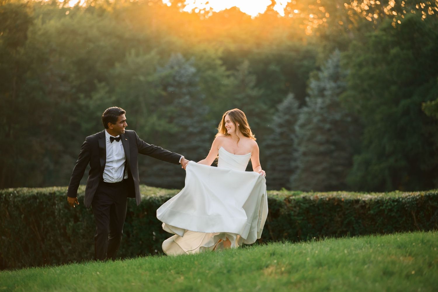 Couple in wedding attire walking at sunset, bride lifting white gown, groom in tuxedo, lush green hedges background.