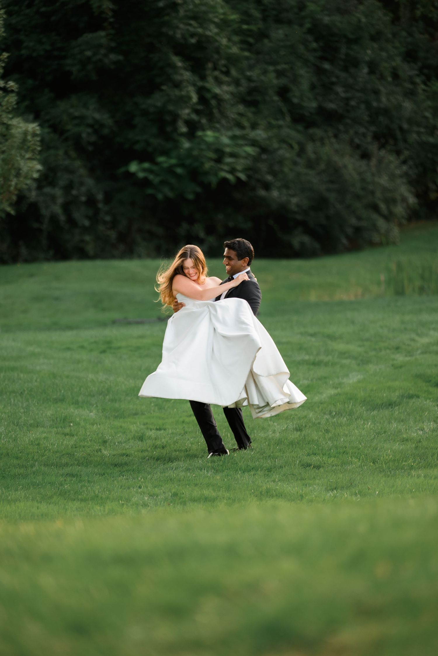 Groom lifting bride in white wedding dress on lush green lawn with trees in background.