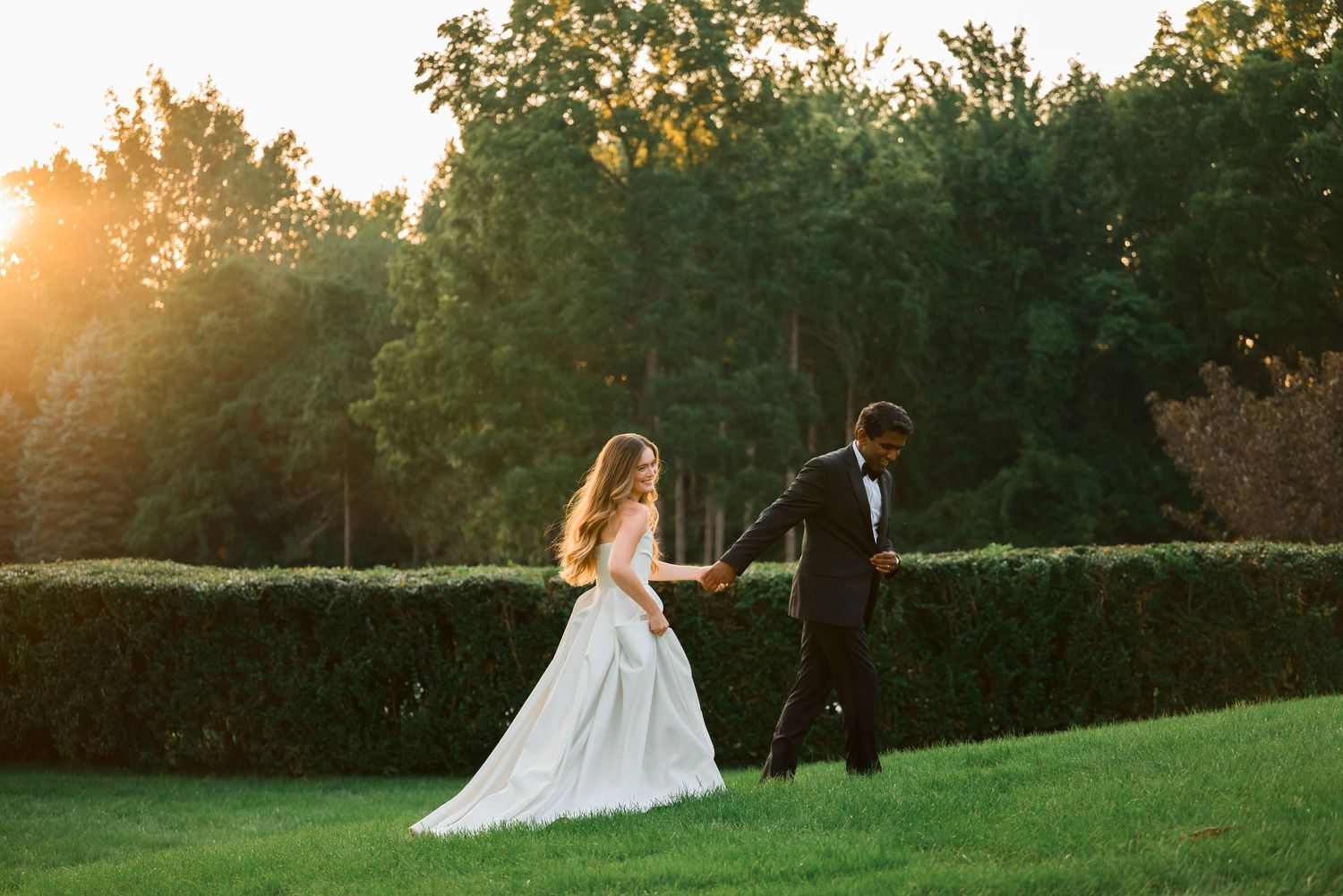 Bride in white gown and groom in tuxedo hold hands walking on green lawn at golden sunset.