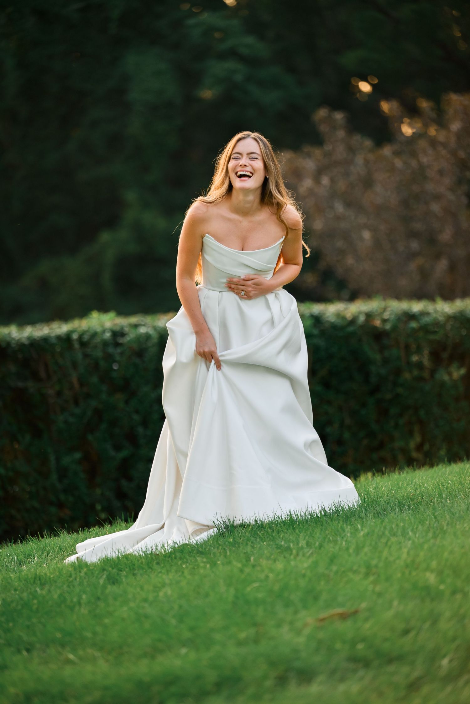 A laughing bride in a strapless white gown walks across lush green grass with hedges in the background.