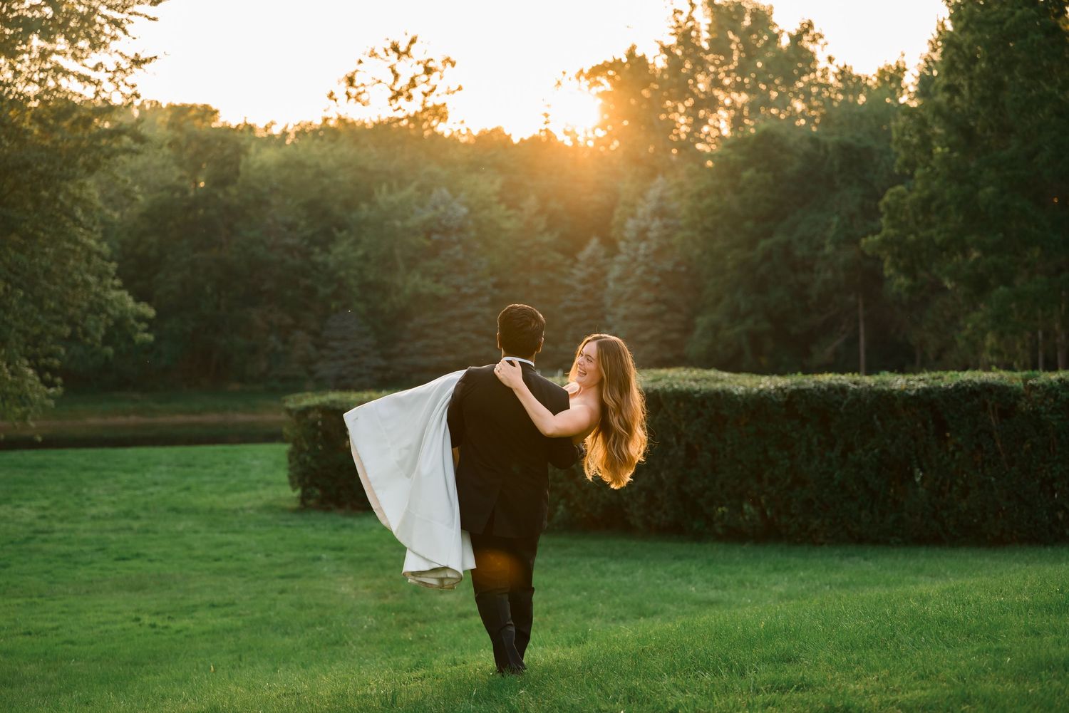 Couple dancing at sunset in lush green garden, bride in white gown lifted by groom in black suit.
