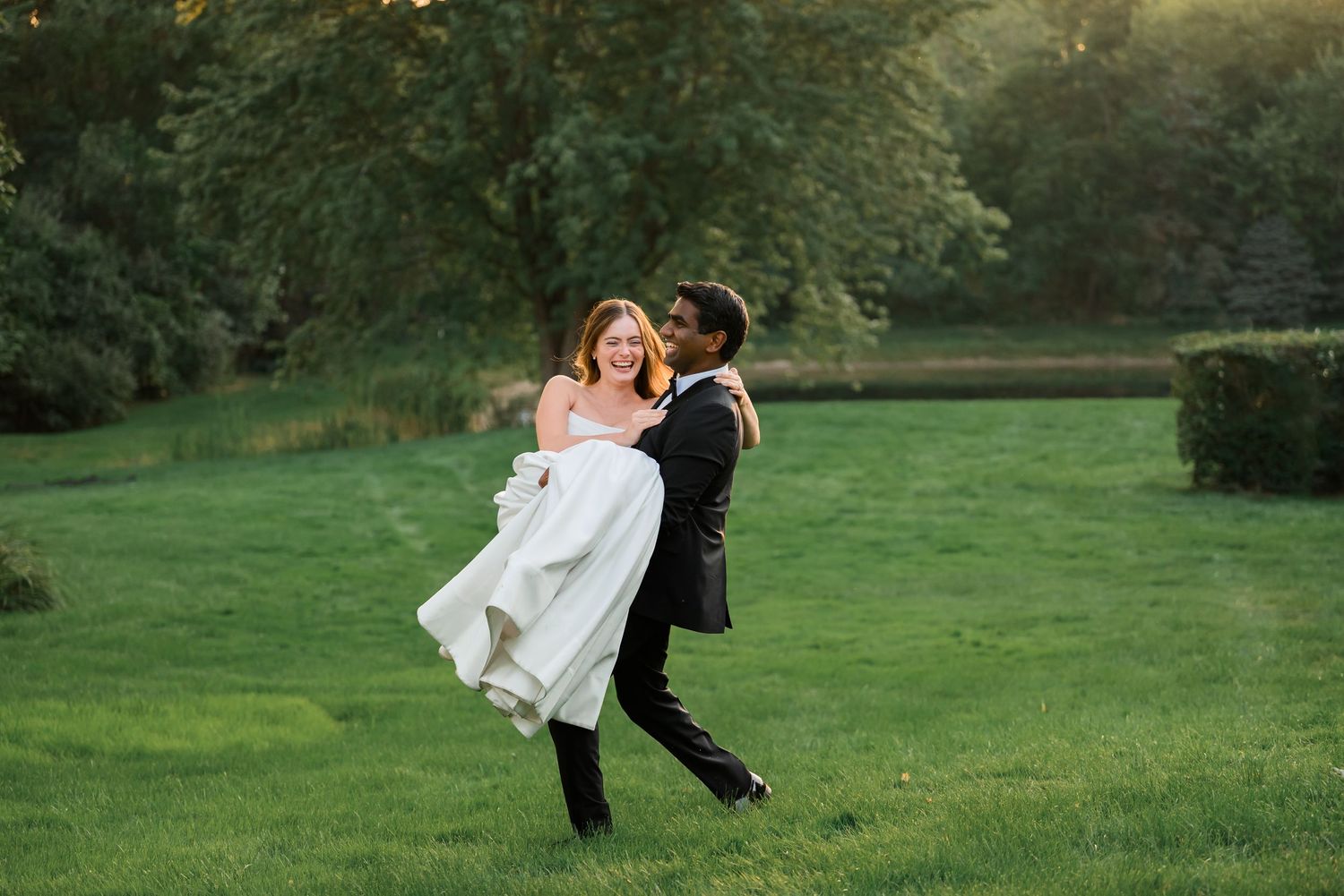 Groom carrying smiling bride in white gown across lush green lawn during golden hour outdoor wedding celebration.