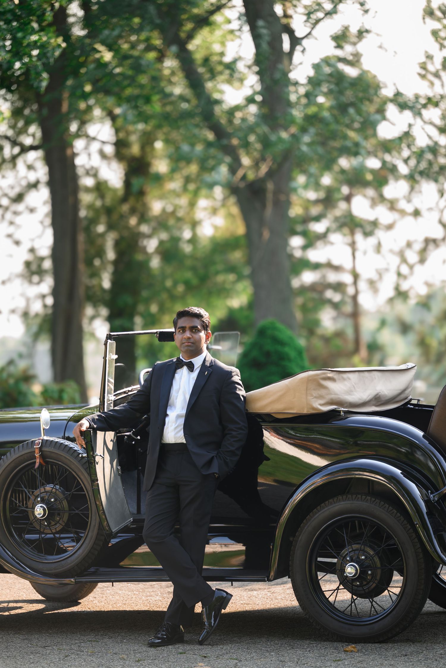 Dapper groom in black tuxedo leaning against vintage classic black car outdoors among trees.