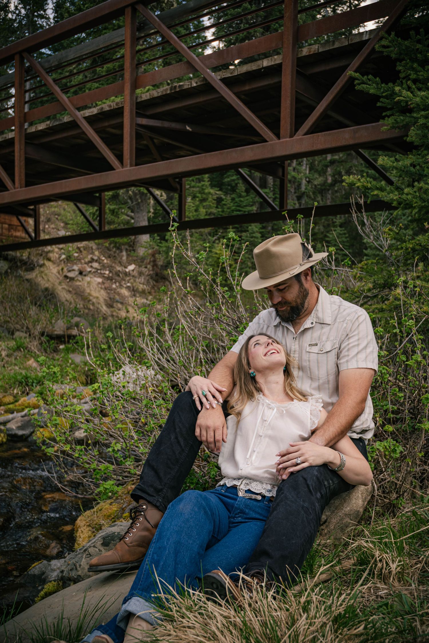 Couple sitting together under a rustic metal bridge in a forest, man in cowboy hat embracing woman in white blouse.