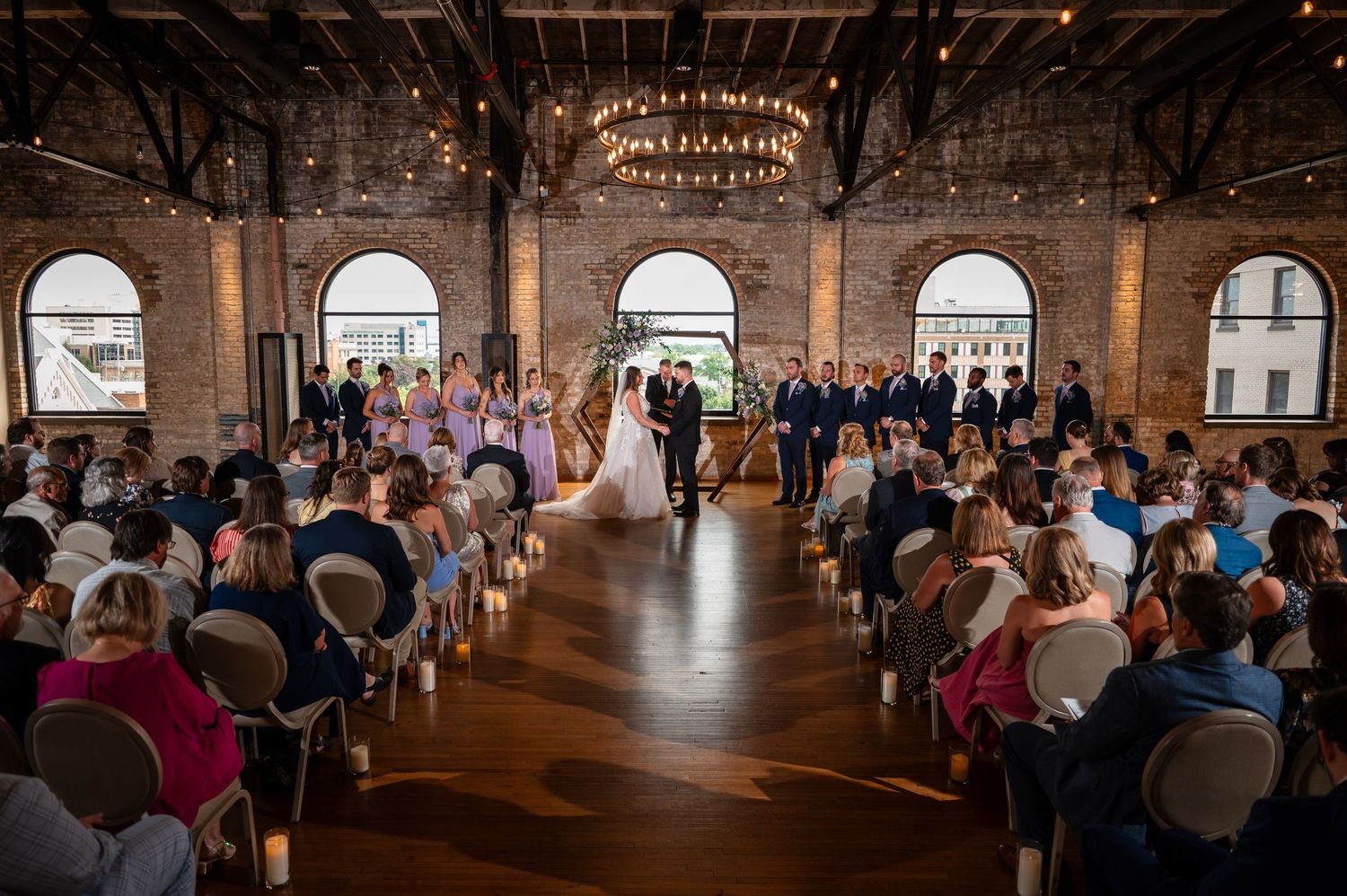 Elegant indoor wedding ceremony in a rustic brick venue with chandeliers, candles, and guests seated as couple stands at altar.