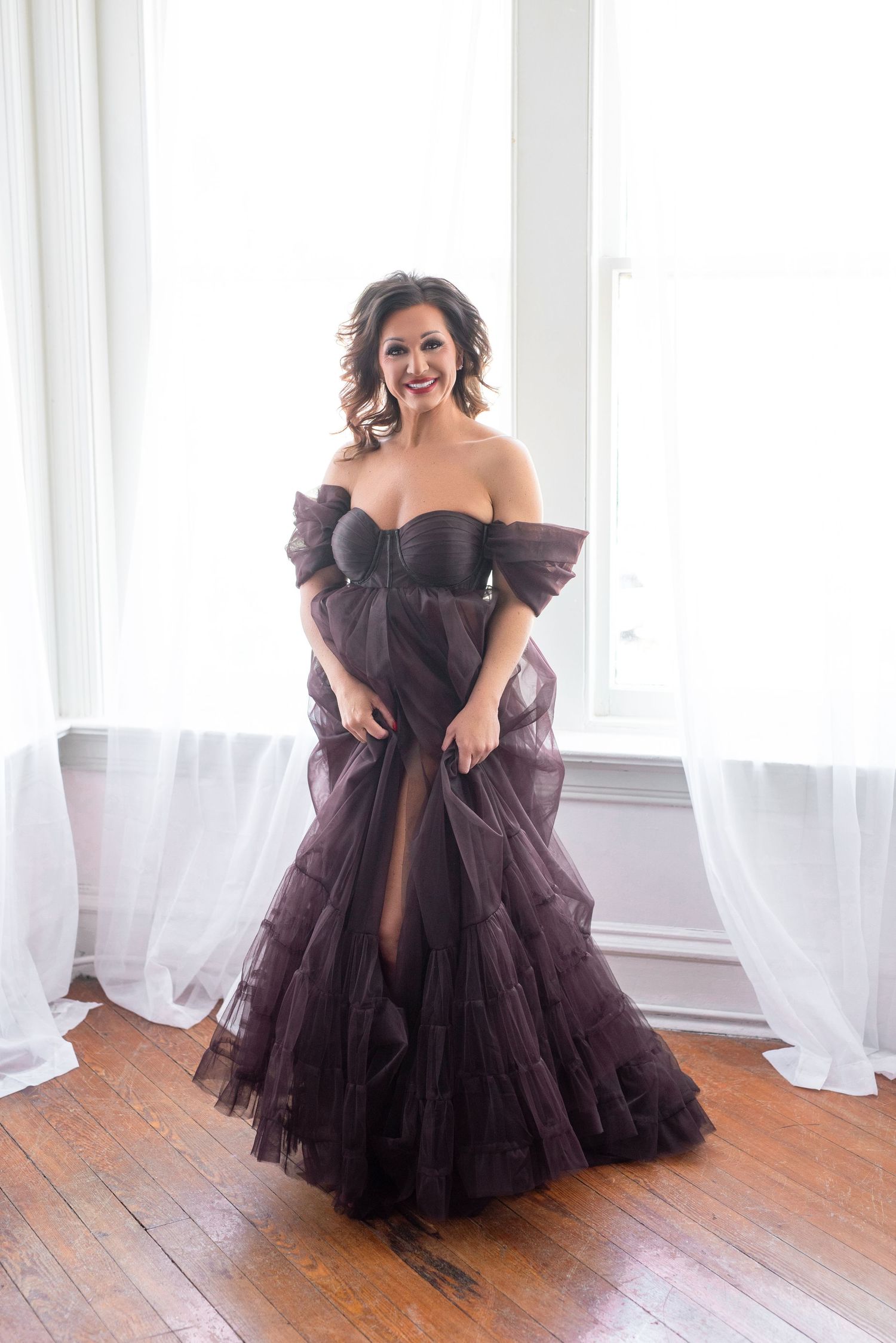 Woman in elegant off-shoulder dark purple tulle gown posing gracefully in bright white room with hardwood floors.