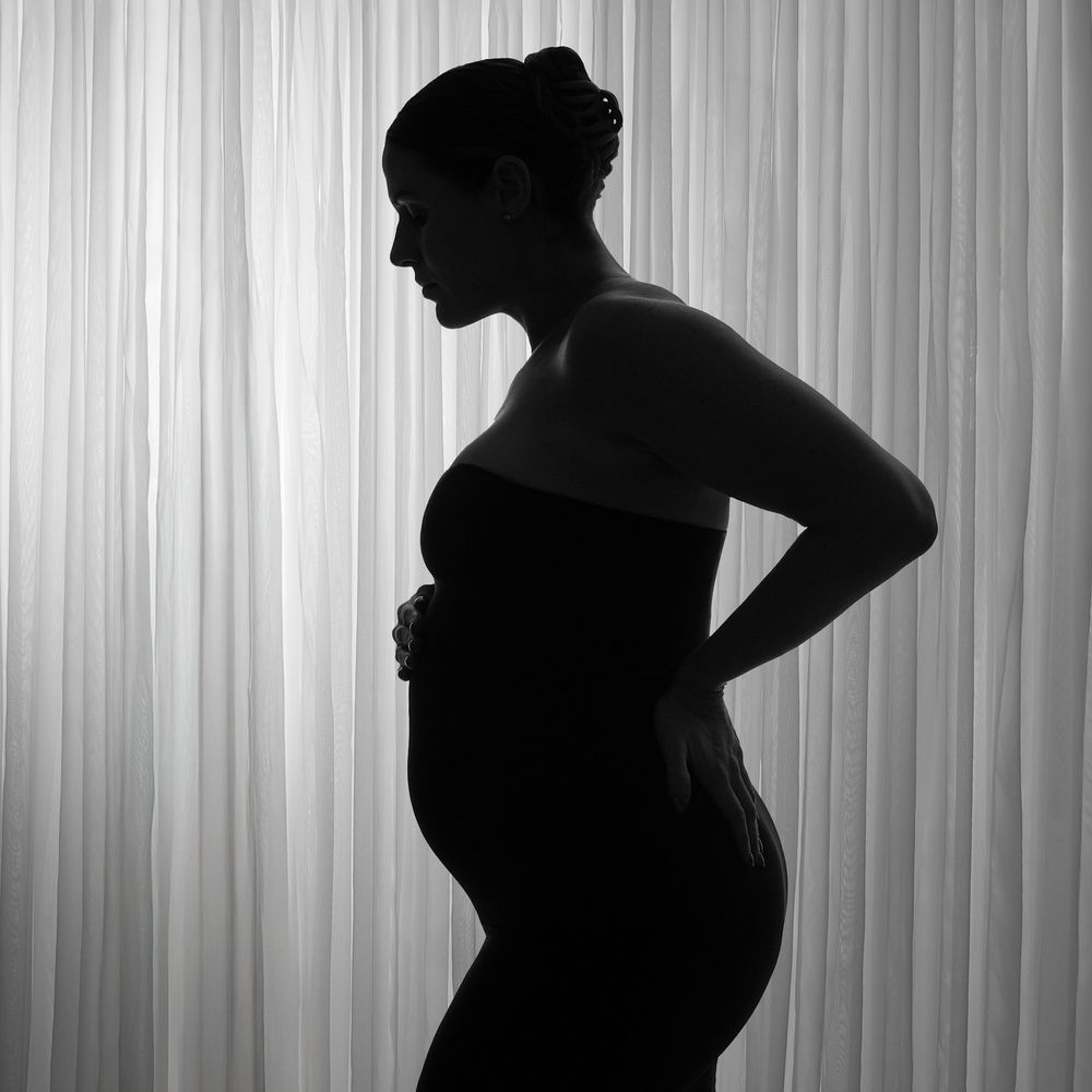 Black and white silhouette of a pregnant woman cradling her baby bump against a soft curtain backdrop.