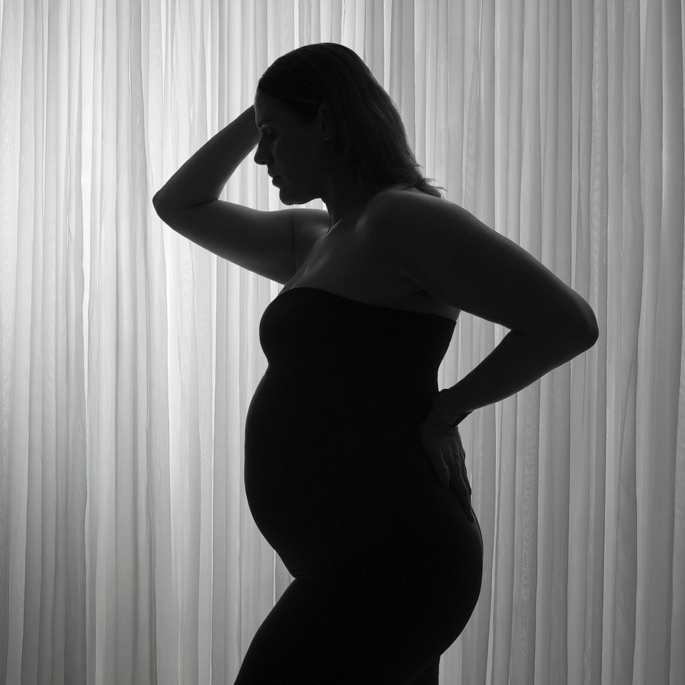 Black and white silhouette of a pregnant woman posing gracefully against a soft curtain backdrop.