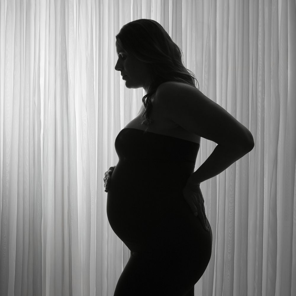 Black and white silhouette of a pregnant woman standing gracefully against a softly lit curtain backdrop.