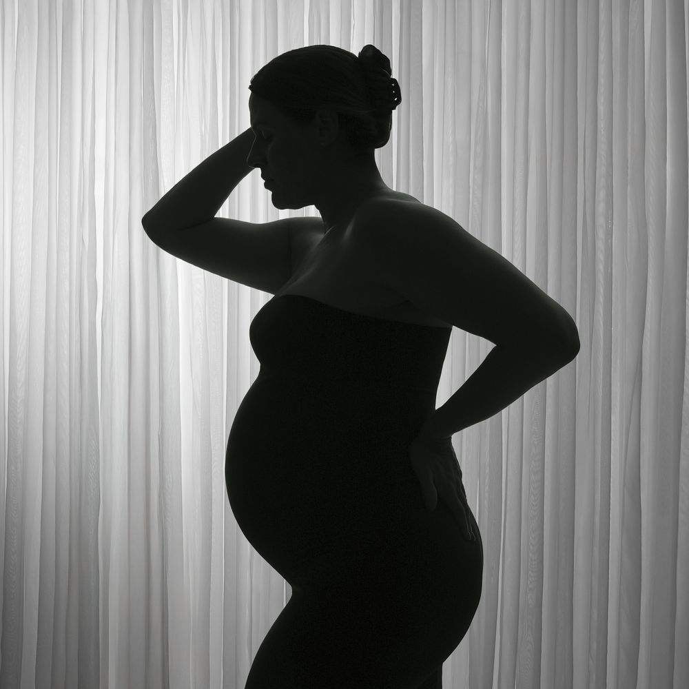 Black and white silhouette of a pregnant woman posing gracefully against a soft draped curtain background.
