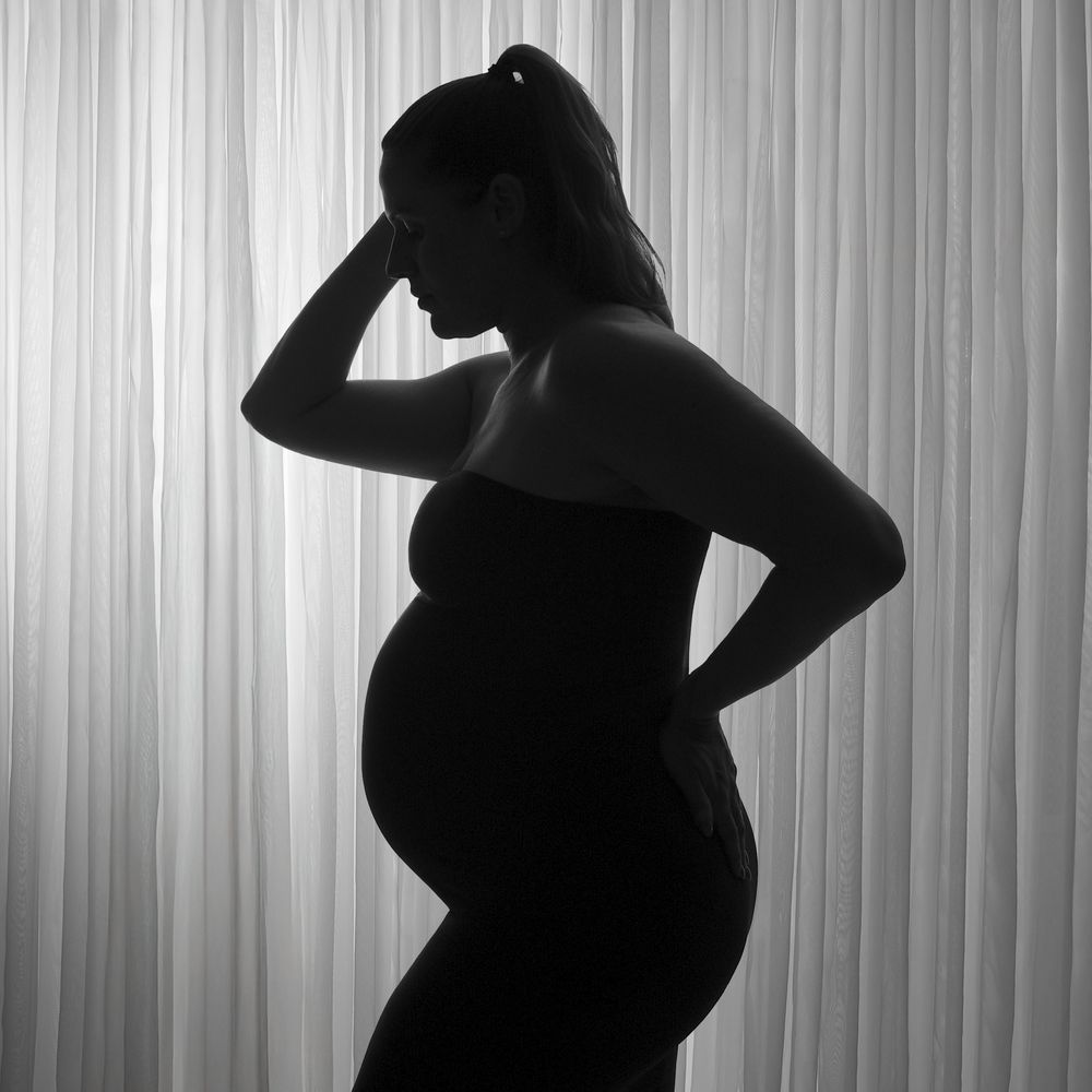 Black and white silhouette of a pregnant woman posing gracefully against a soft curtain backdrop.