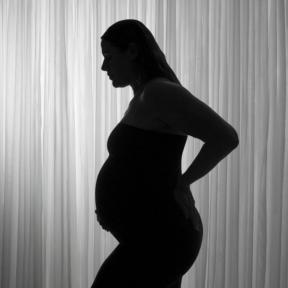 Black and white silhouette of a pregnant woman standing sideways against a white curtain backdrop.