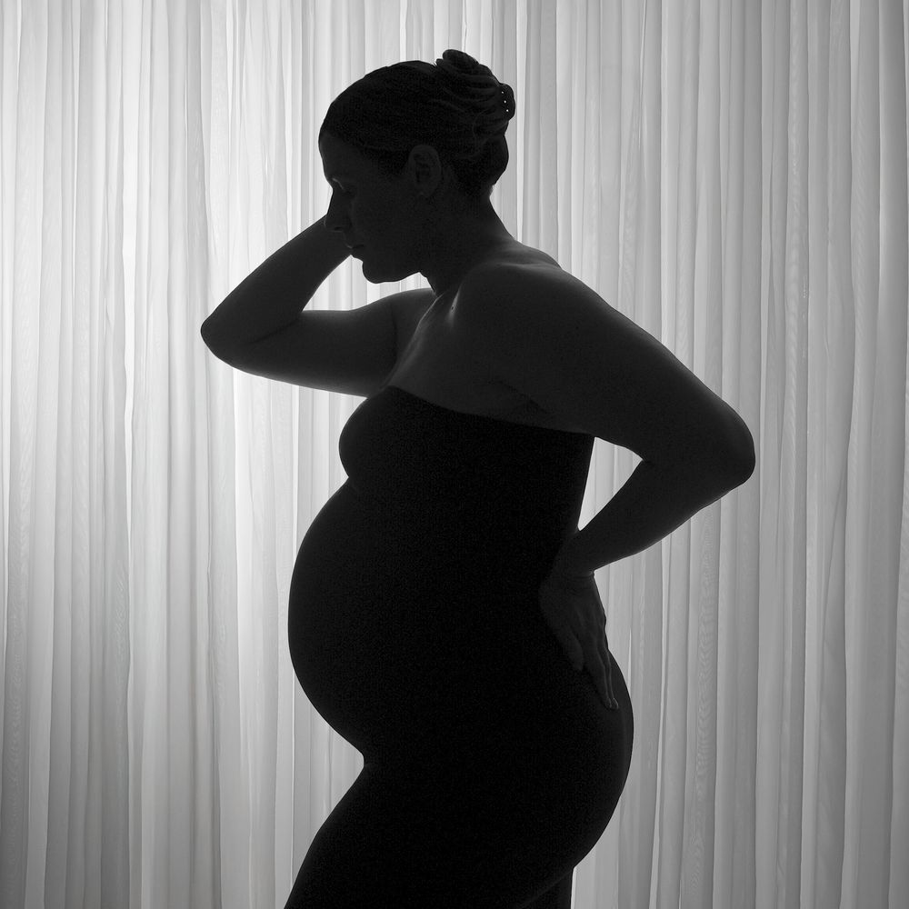 Black and white silhouette of a pregnant woman standing gracefully against a soft curtain backdrop.