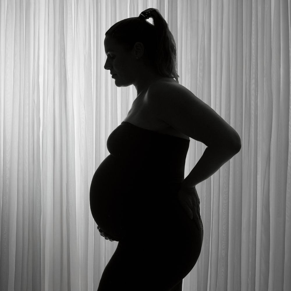 Black and white silhouette of a pregnant woman standing sideways against sheer curtains, showcasing her baby bump.