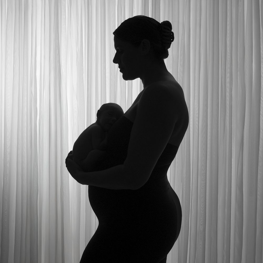 Black and white silhouette of a pregnant woman holding a teddy bear against a soft curtain backdrop.