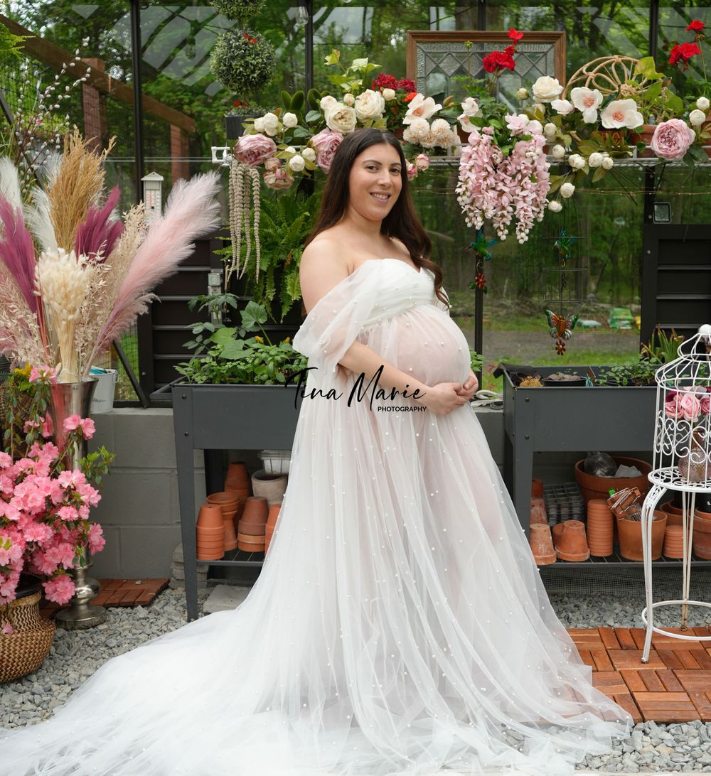 Ethereal maternity photo in flowing white gown surrounded by greenhouse flowers and pink decor.