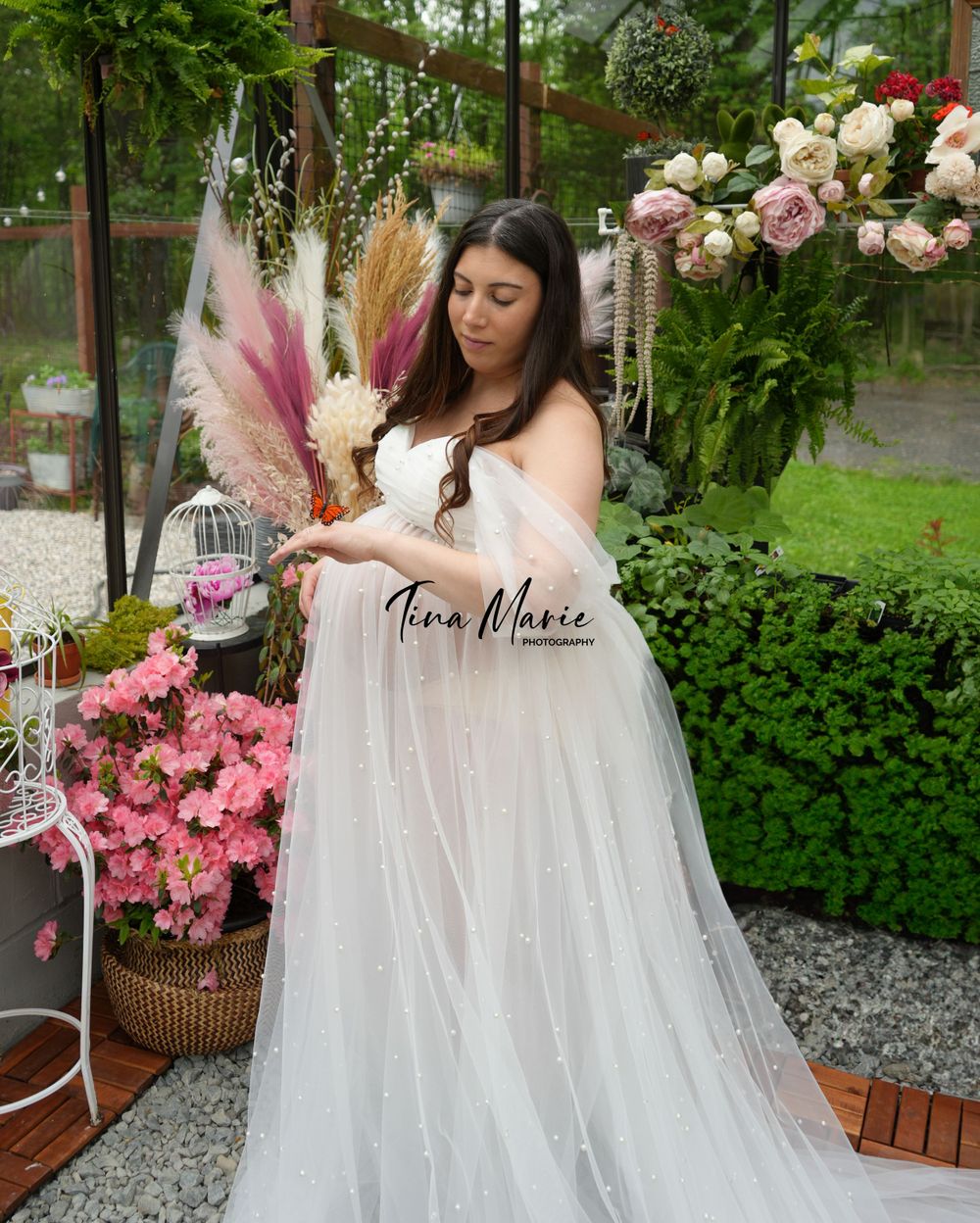 Elegant maternity portrait in white tulle gown with pink florals in greenhouse setting.