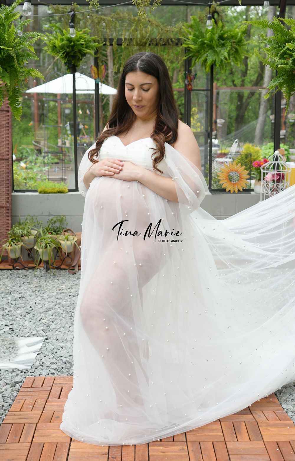 Ethereal maternity portrait with a flowing white dress in a garden greenhouse setting.