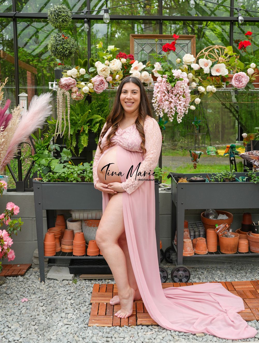Greenhouse maternity photo with pink dress surrounded by roses and garden decor.