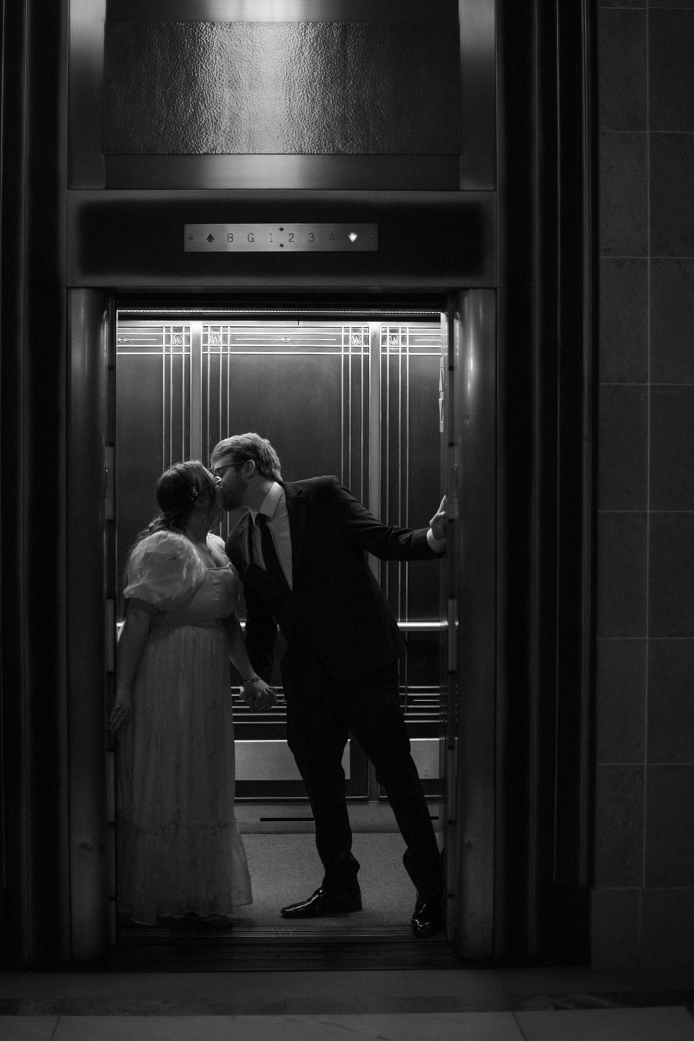 A bride and groom share a romantic kiss inside an elegant elevator in this black and white wedding photo.
