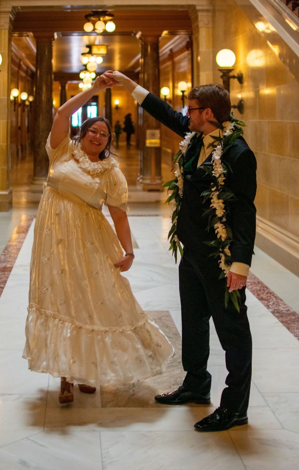 Couple dancing in elegant hallway, bride in vintage white gown, groom in black suit with floral lei.