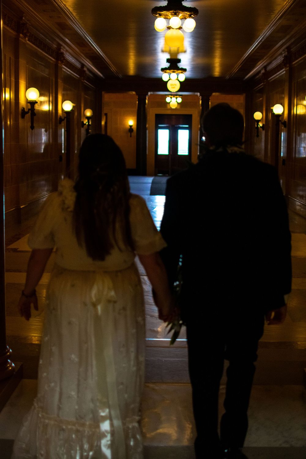 Bride and groom walking hand-in-hand down a warmly lit ornate hallway after wedding ceremony.