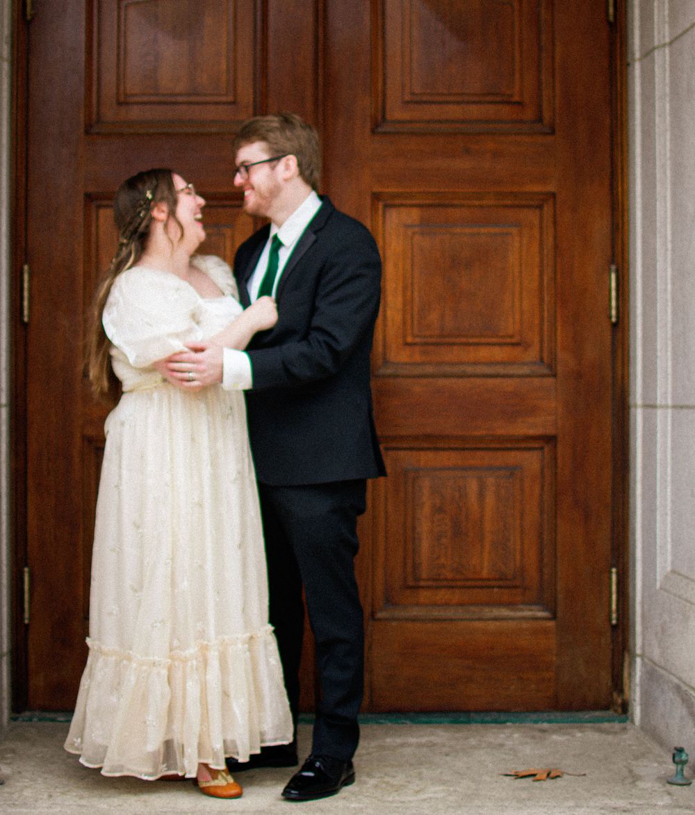 A bride and groom share a tender embrace in front of large ornate wooden doors at their wedding venue.