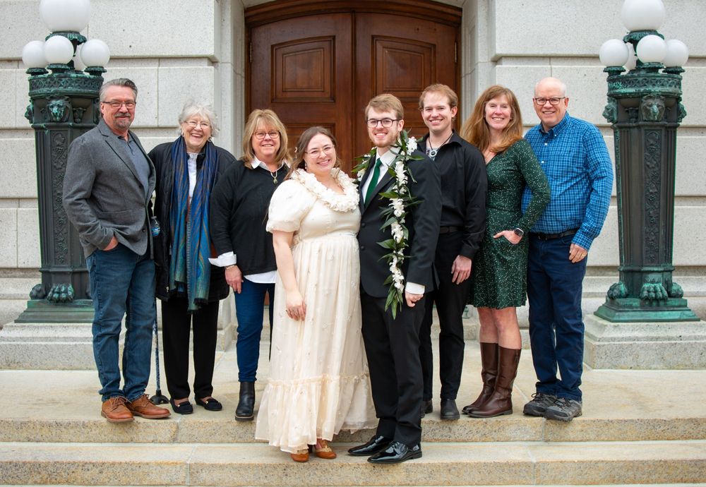 Wedding party group photo on building steps, bride in white dress with groom wearing floral lei.