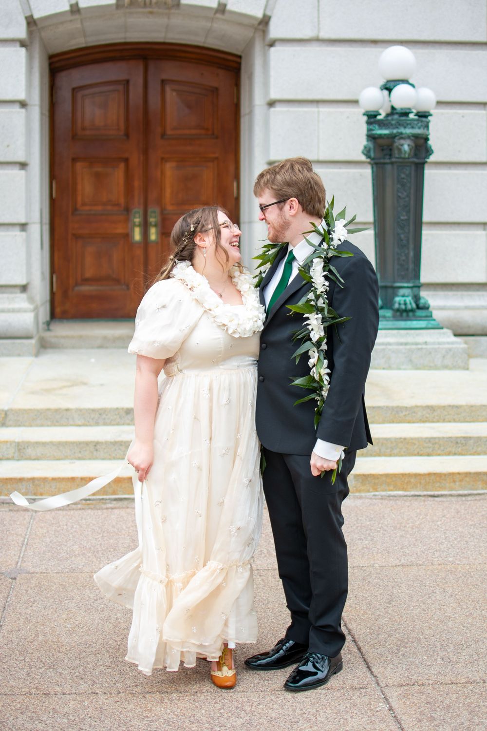 A laughing bride and groom share a joyful moment outside a grand stone building with wooden doors.