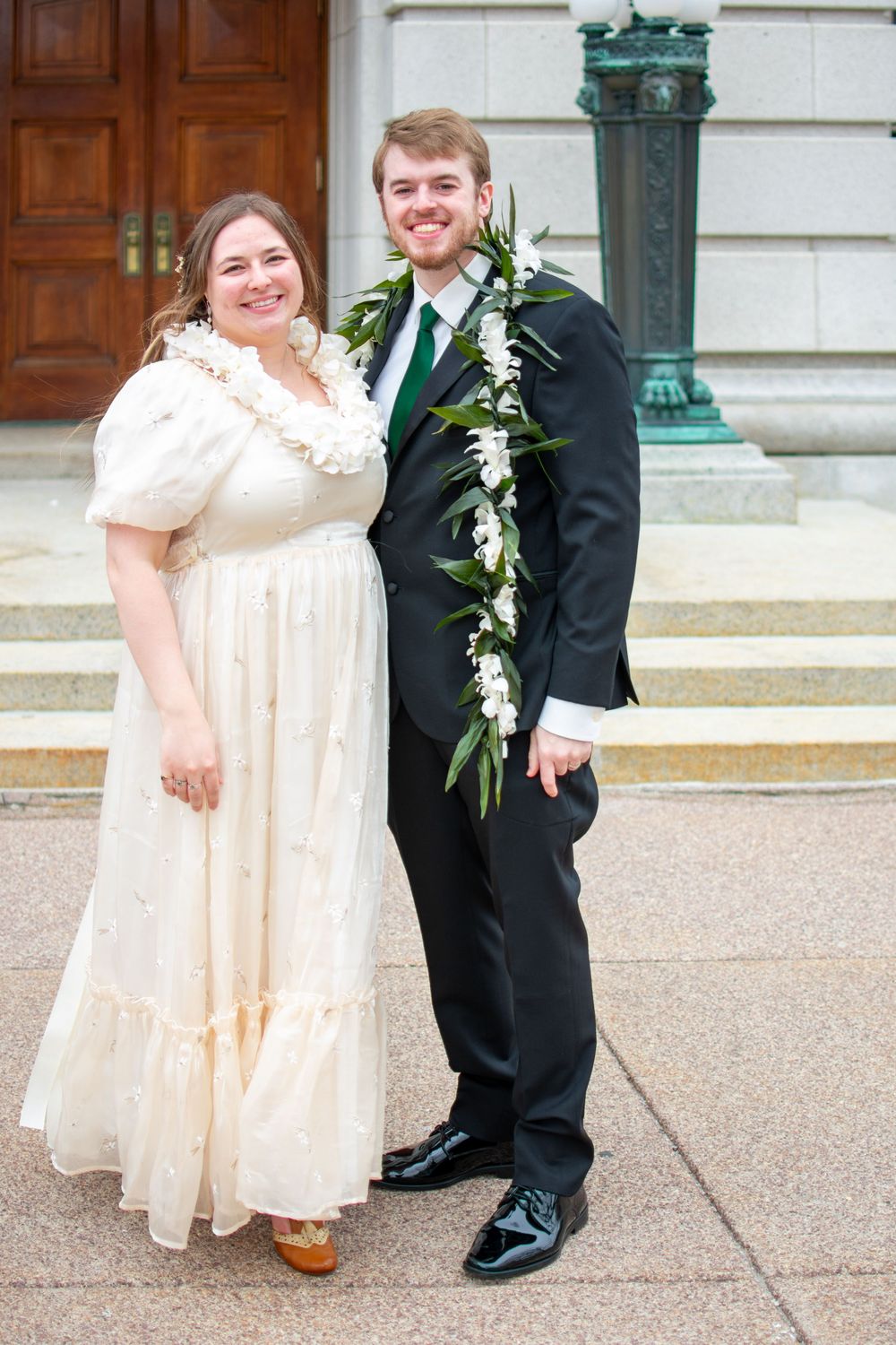 A smiling bride and groom pose outdoors wearing floral leis in front of a grand stone building.