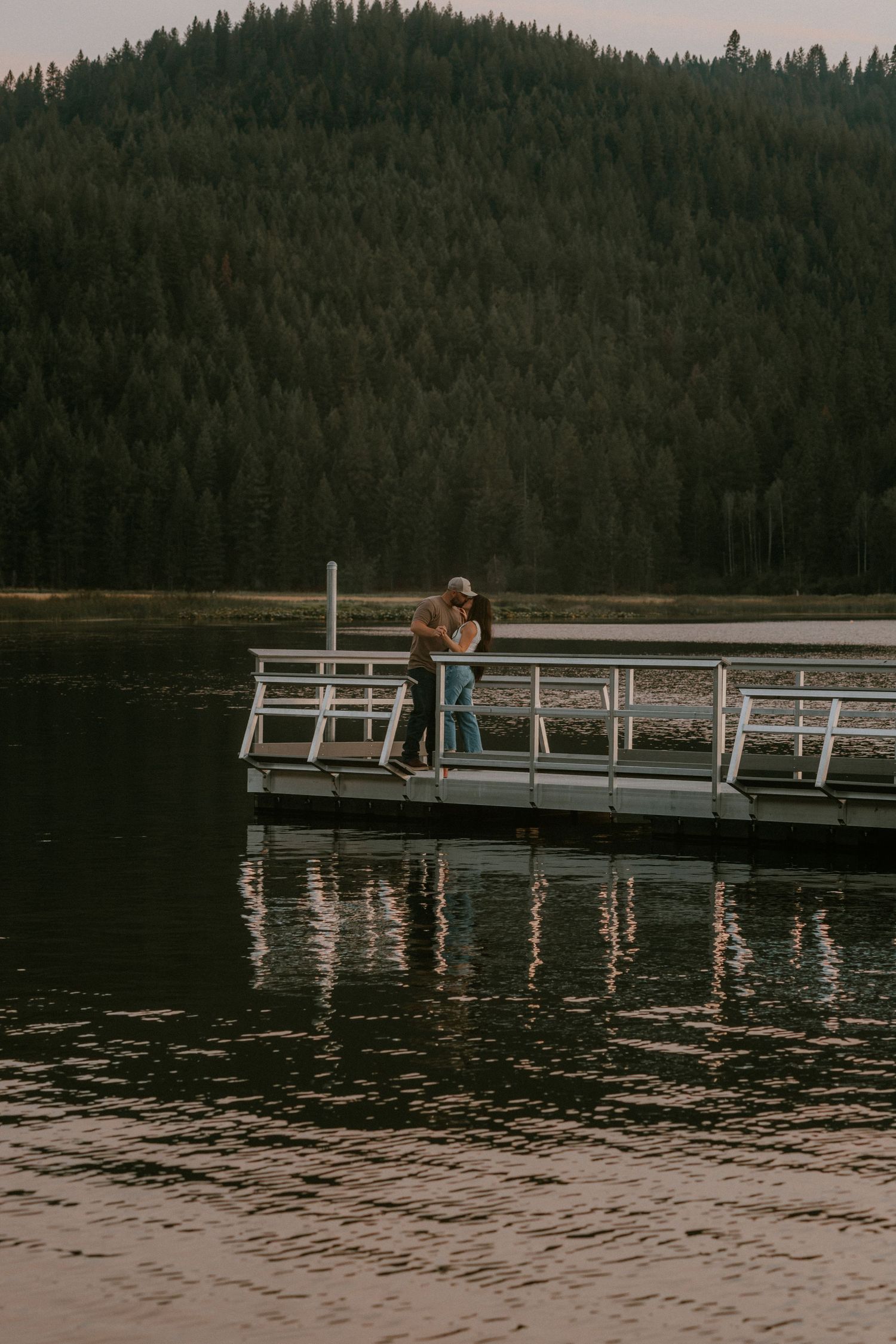 Couple embracing on a wooden dock over a calm lake with a dense evergreen forest and mountain backdrop.