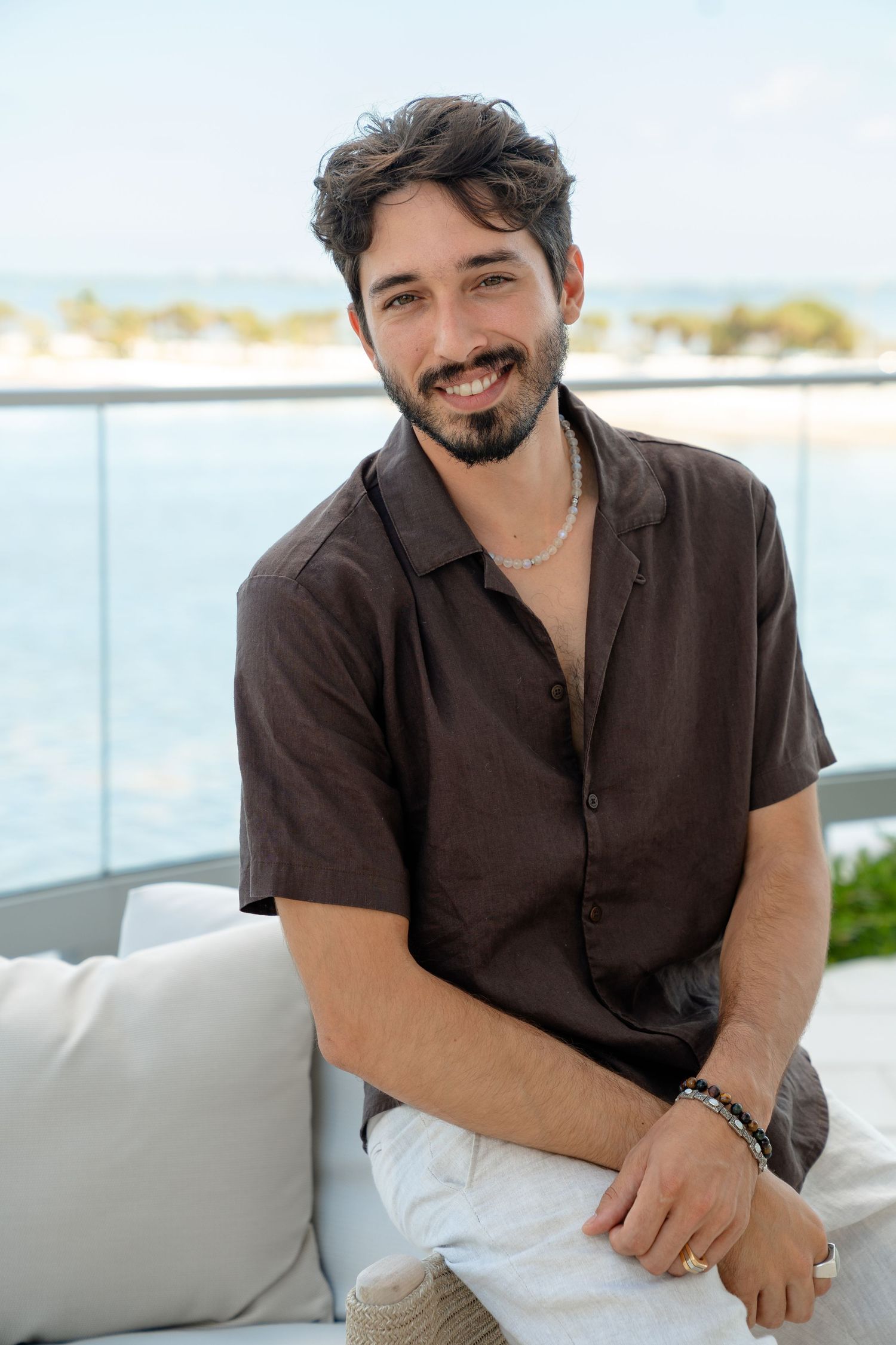Smiling young man with beard wearing brown shirt and necklace sitting on outdoor sofa with ocean view.