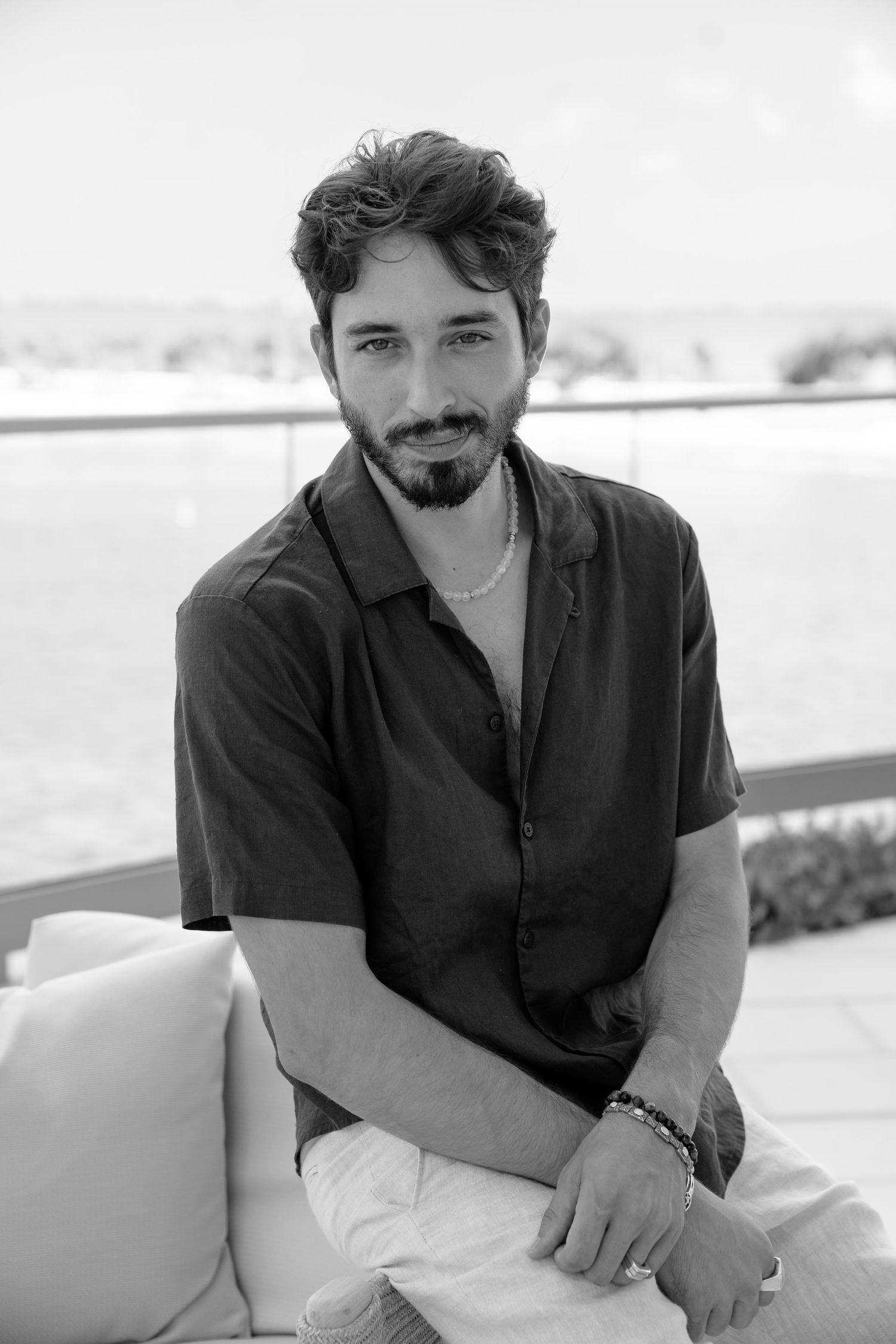 Black and white portrait of a young bearded man sitting on outdoor sofa wearing a dark shirt and necklace.