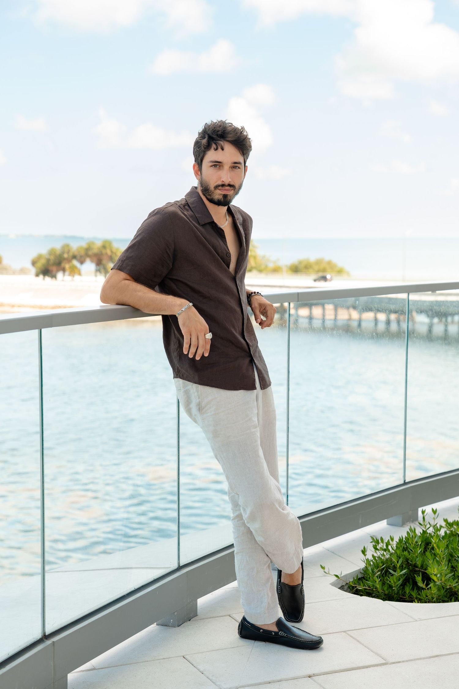 A stylish man in a brown shirt and light pants leans on a glass railing overlooking a turquoise ocean.
