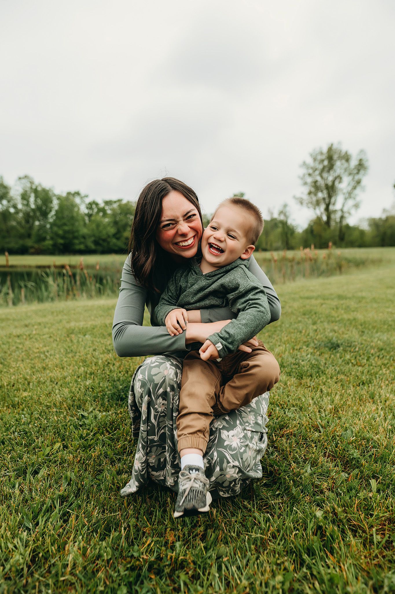 A laughing mother hugs her joyful toddler son while sitting on grass in a lush green park.