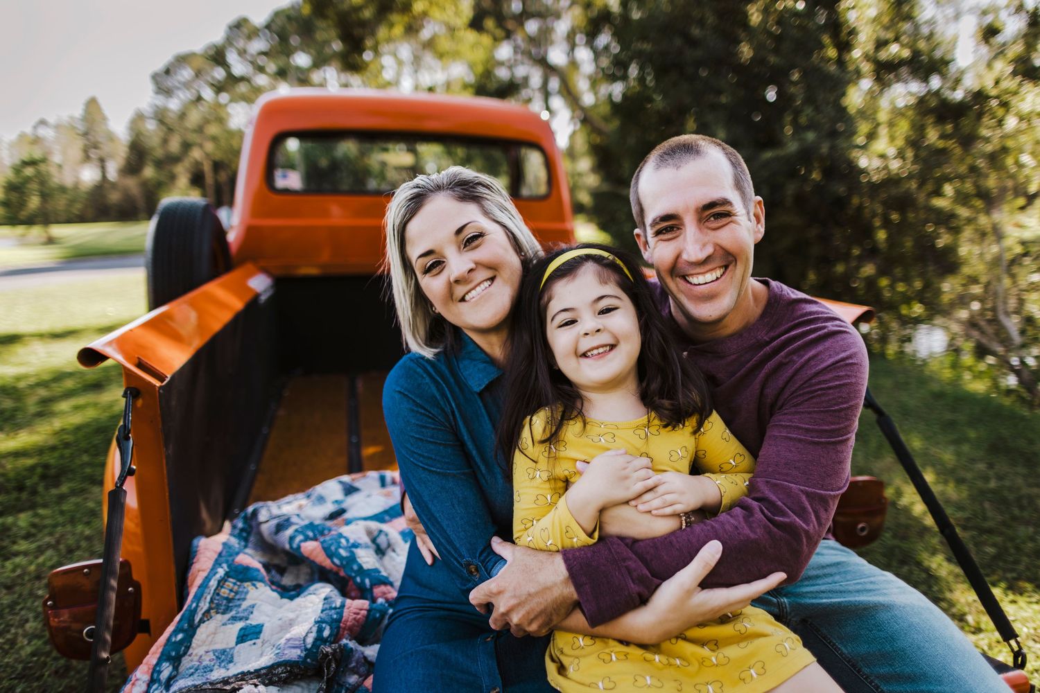 Family of three smiling and hugging in truck bed outdoors.