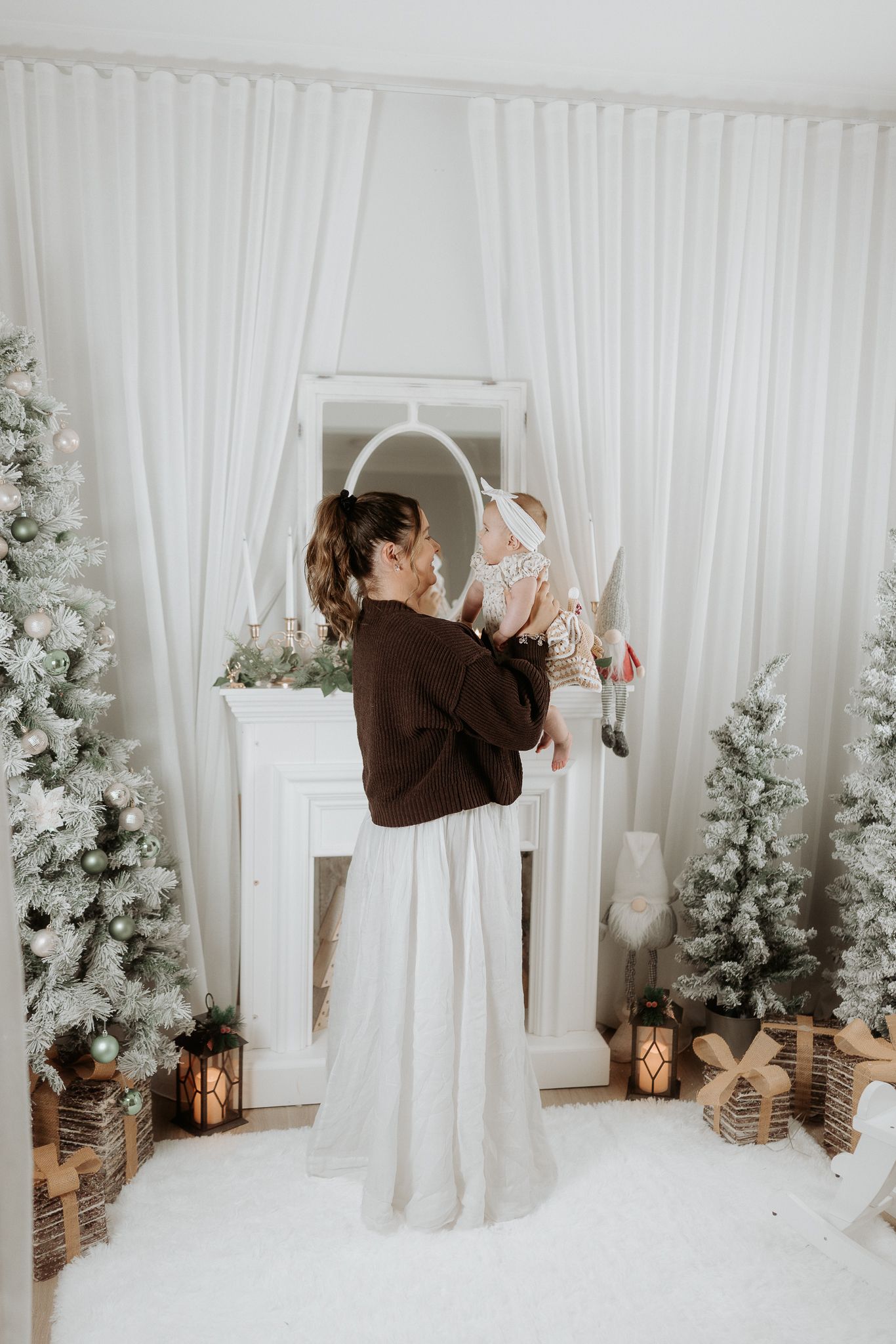 Mother lifting baby near white fireplace with snow-flocked Christmas trees and warm lanterns in festive holiday decor.