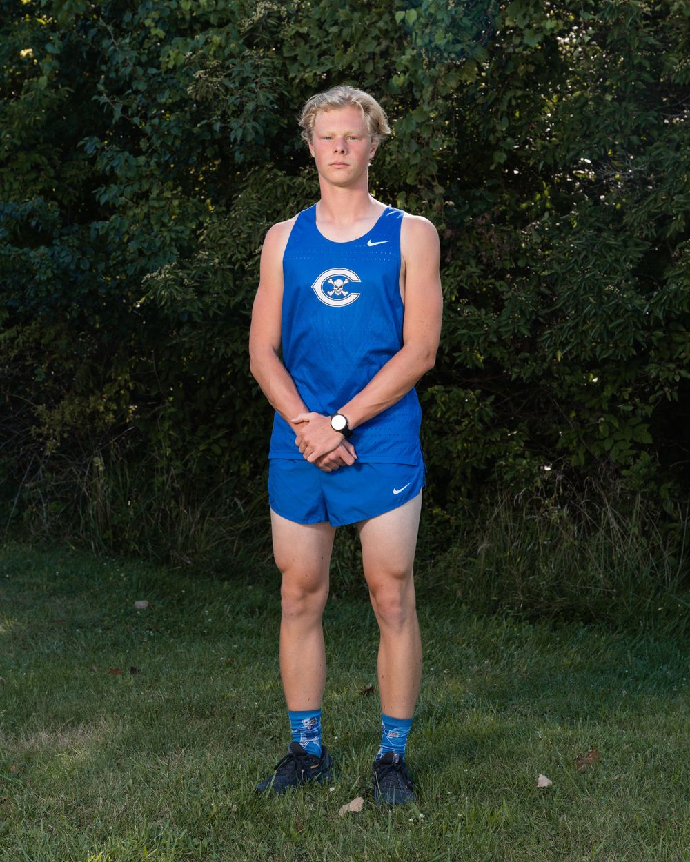 Young male cross country runner in blue Nike uniform stands on grass before green trees.