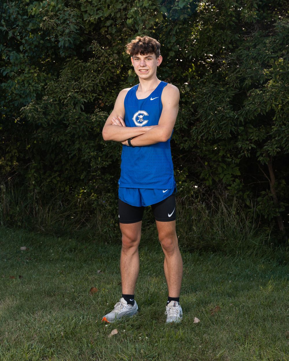 Young male cross country runner in blue Nike uniform stands confidently with arms crossed on green grass.