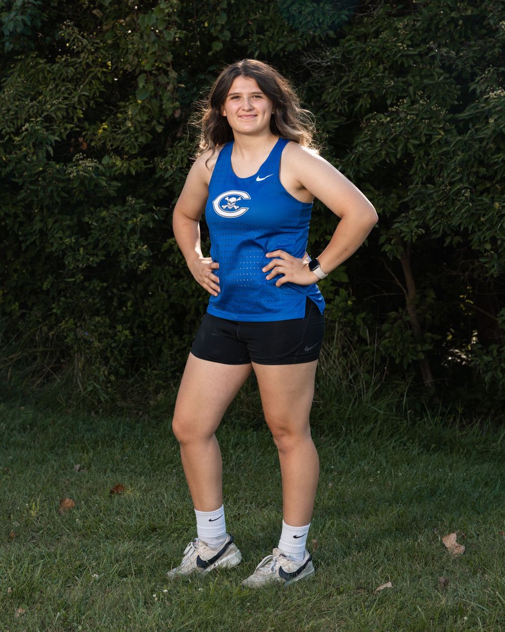 Female athlete in blue Nike track uniform poses confidently on grass with hands on hips during team photo session.