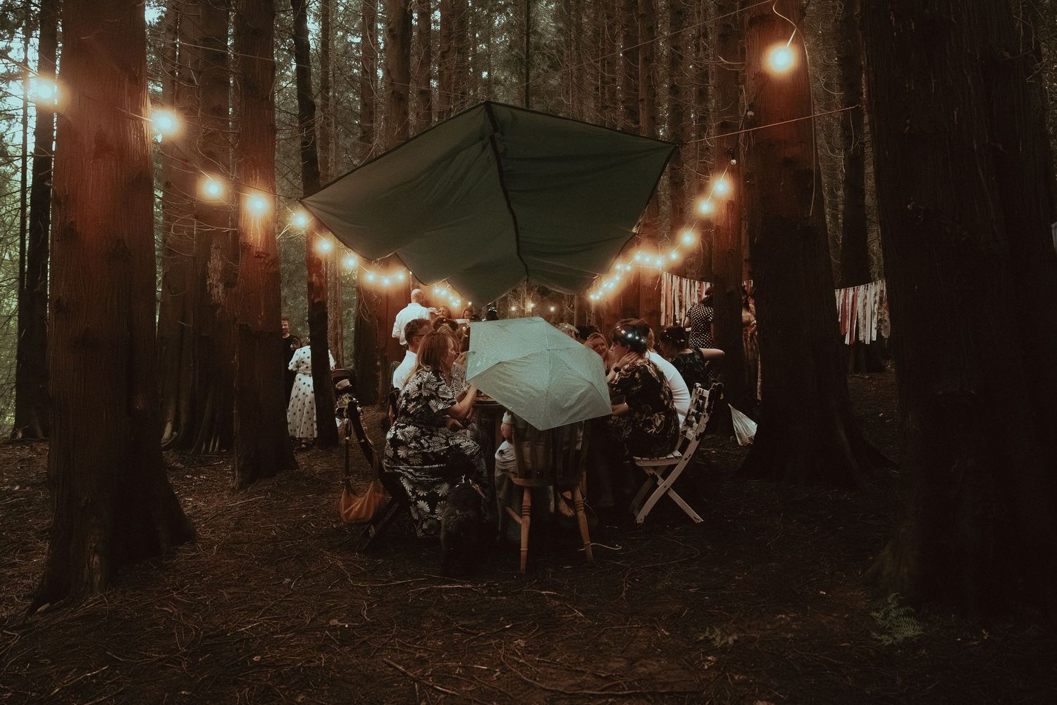 People gathered under a green tarp with string lights in a dark forest setting for an outdoor event.