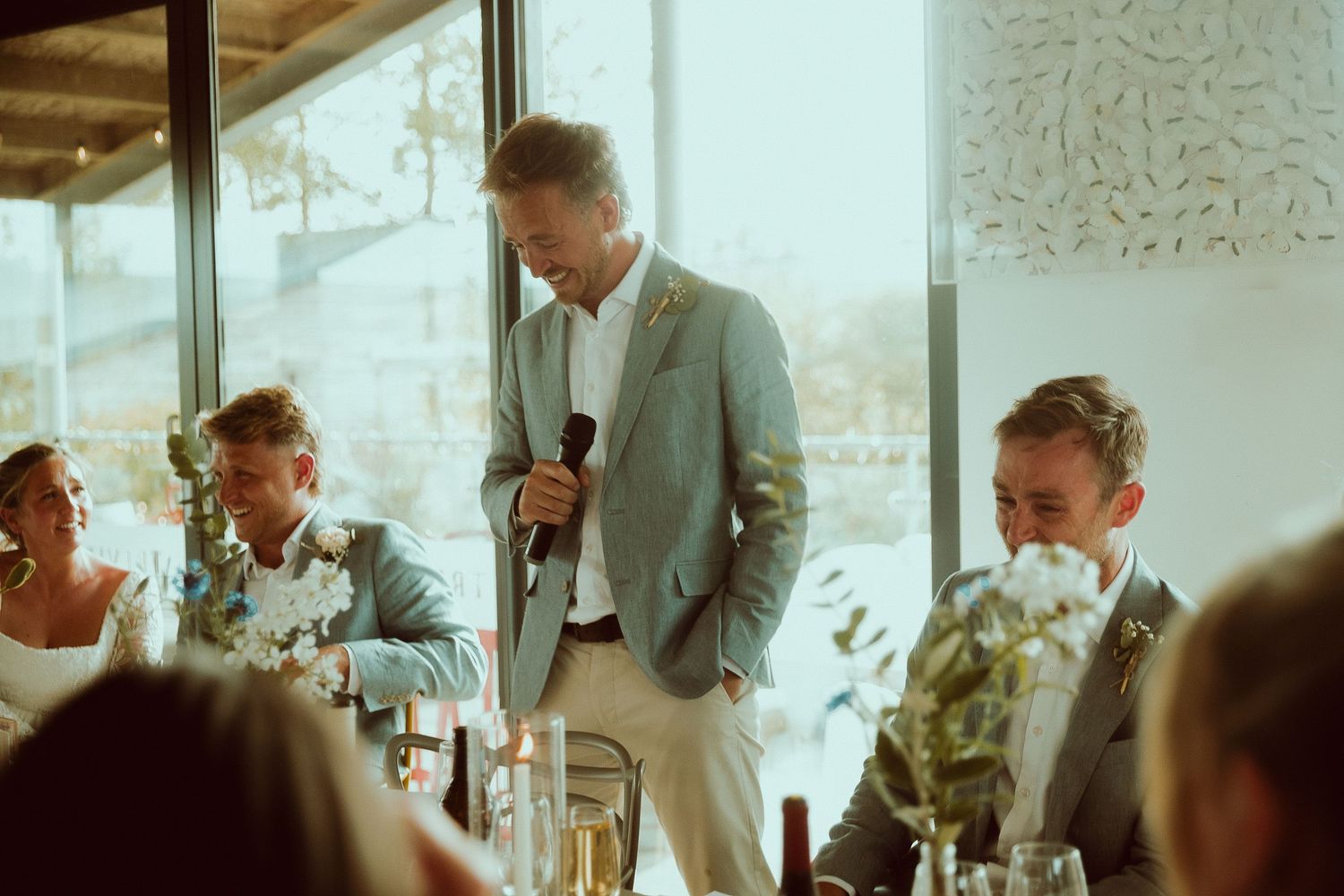 Man in gray suit giving a speech with microphone at an elegant wedding reception dinner.