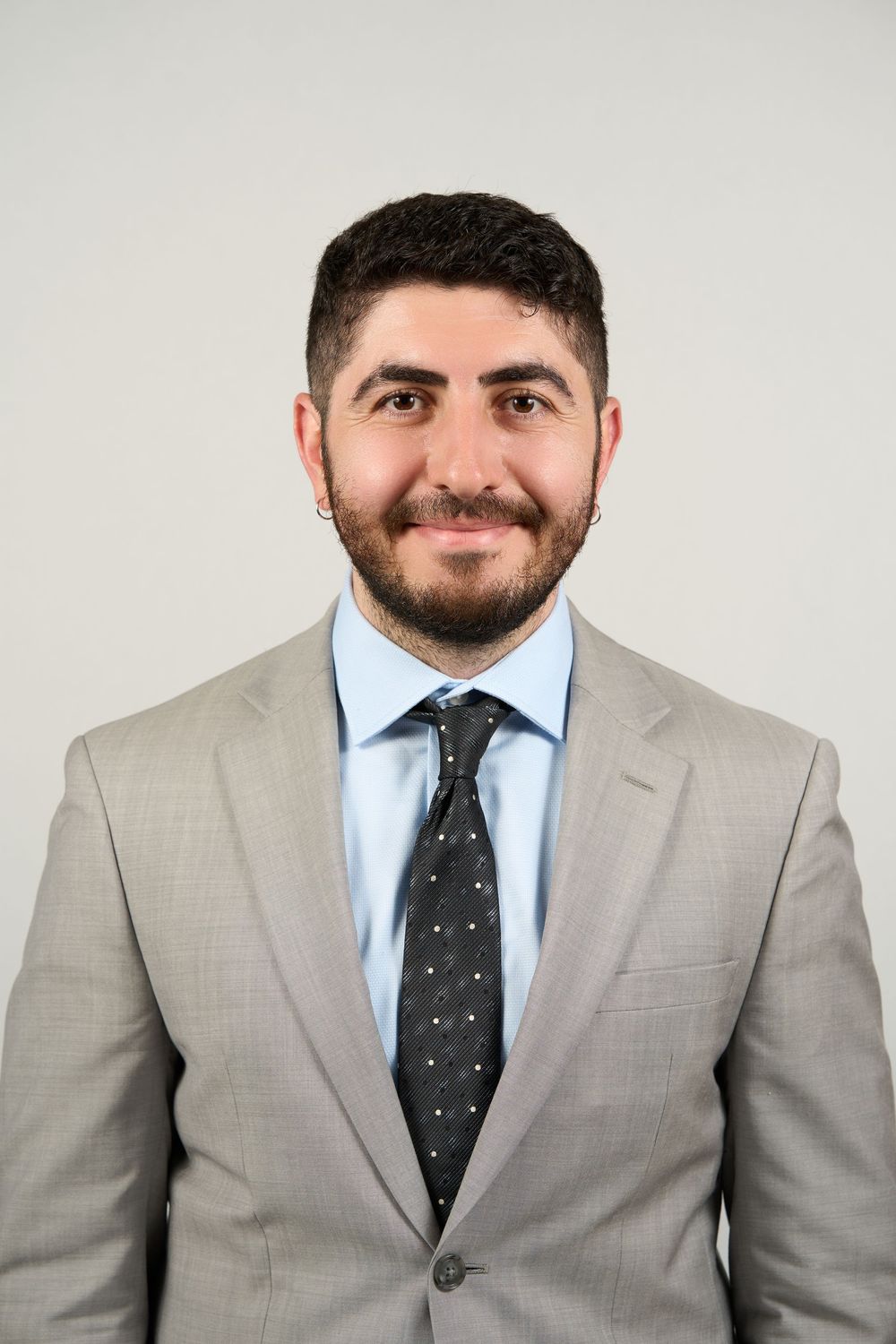 Professional headshot of a smiling young man with beard wearing a light gray suit and dark polka dot tie.