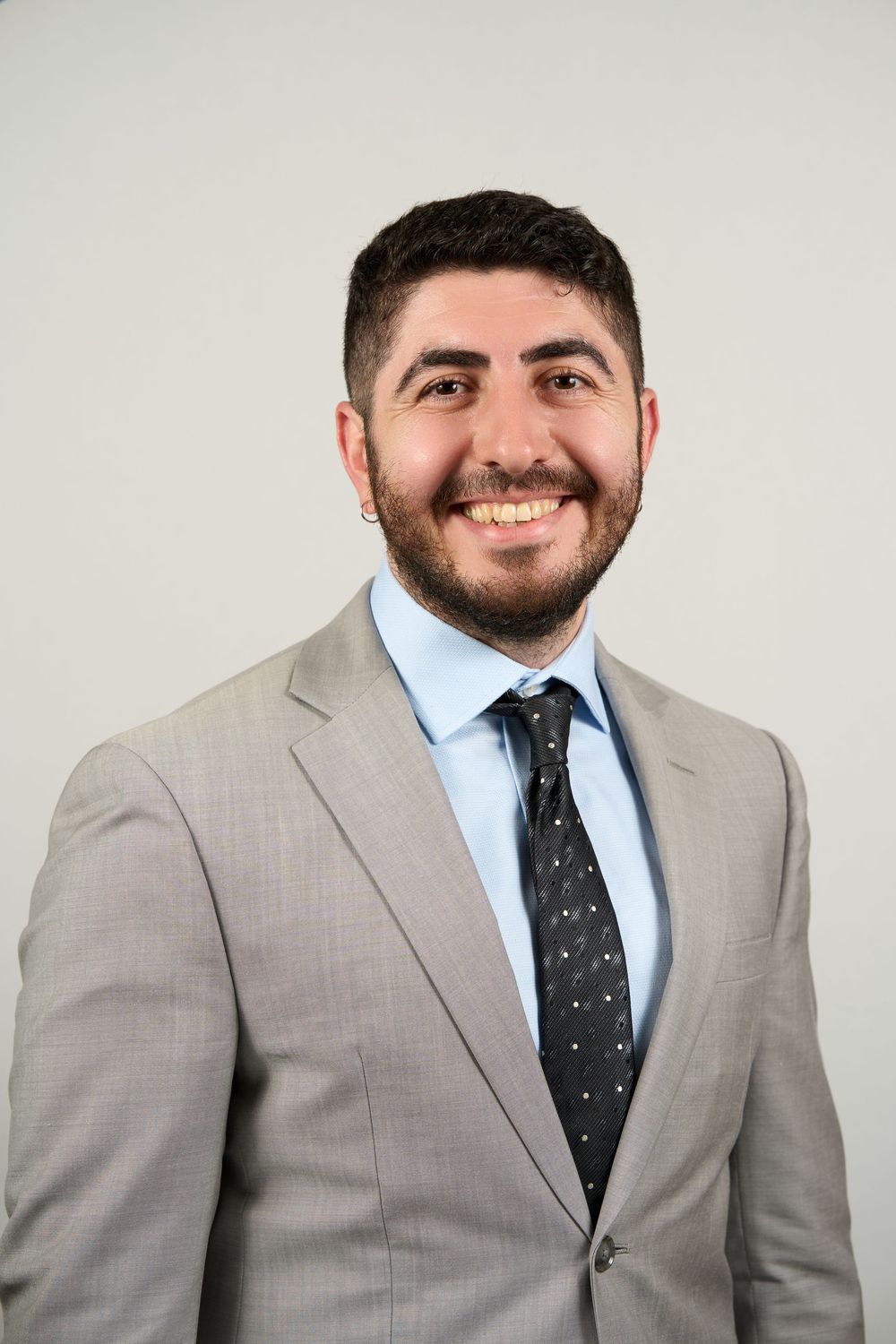 Smiling young man with beard wearing light gray suit, blue dress shirt, and dark dotted tie against gray background.