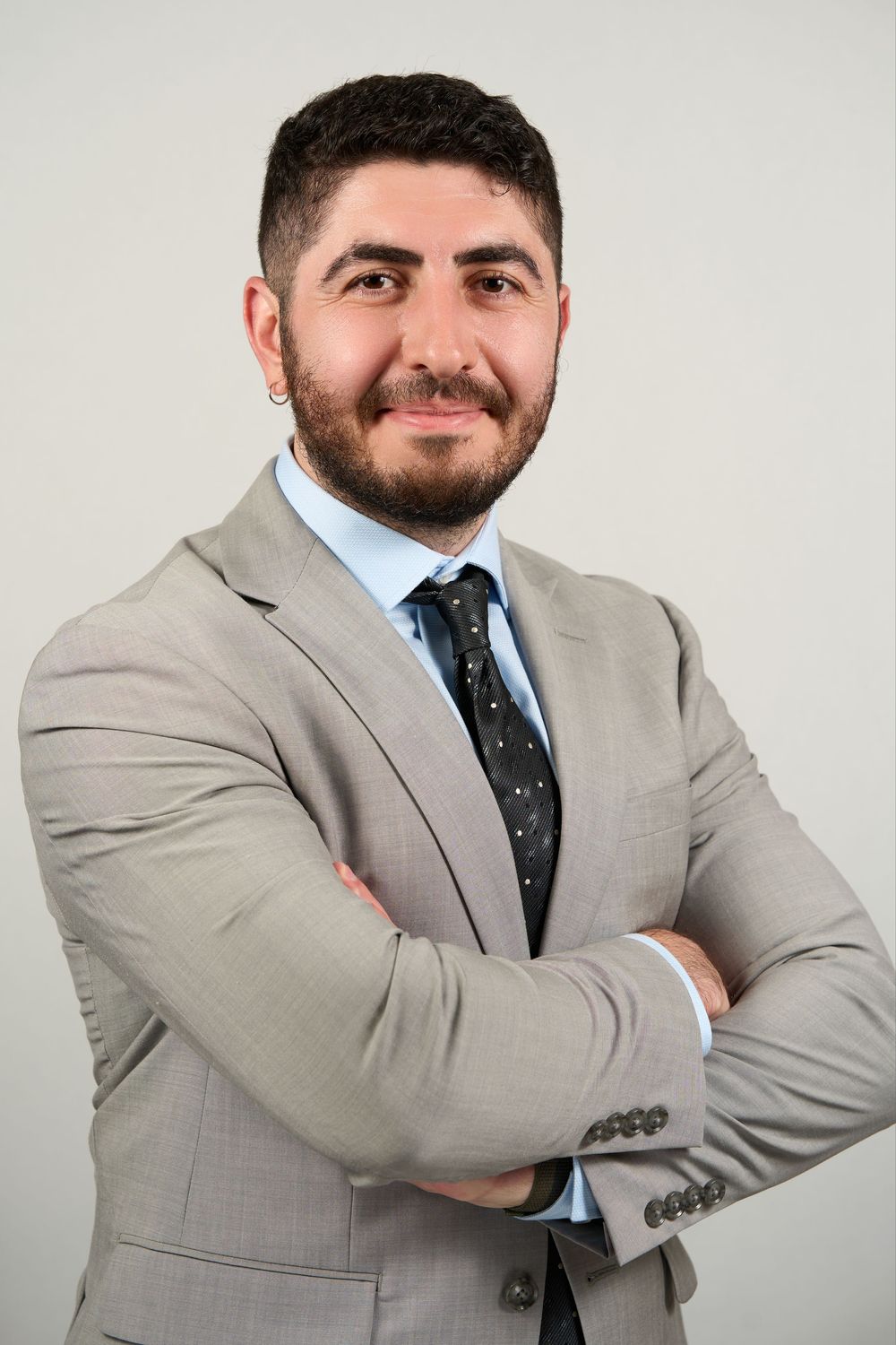 Professional man in gray suit and dark tie with arms crossed, smiling confidently against a light background.
