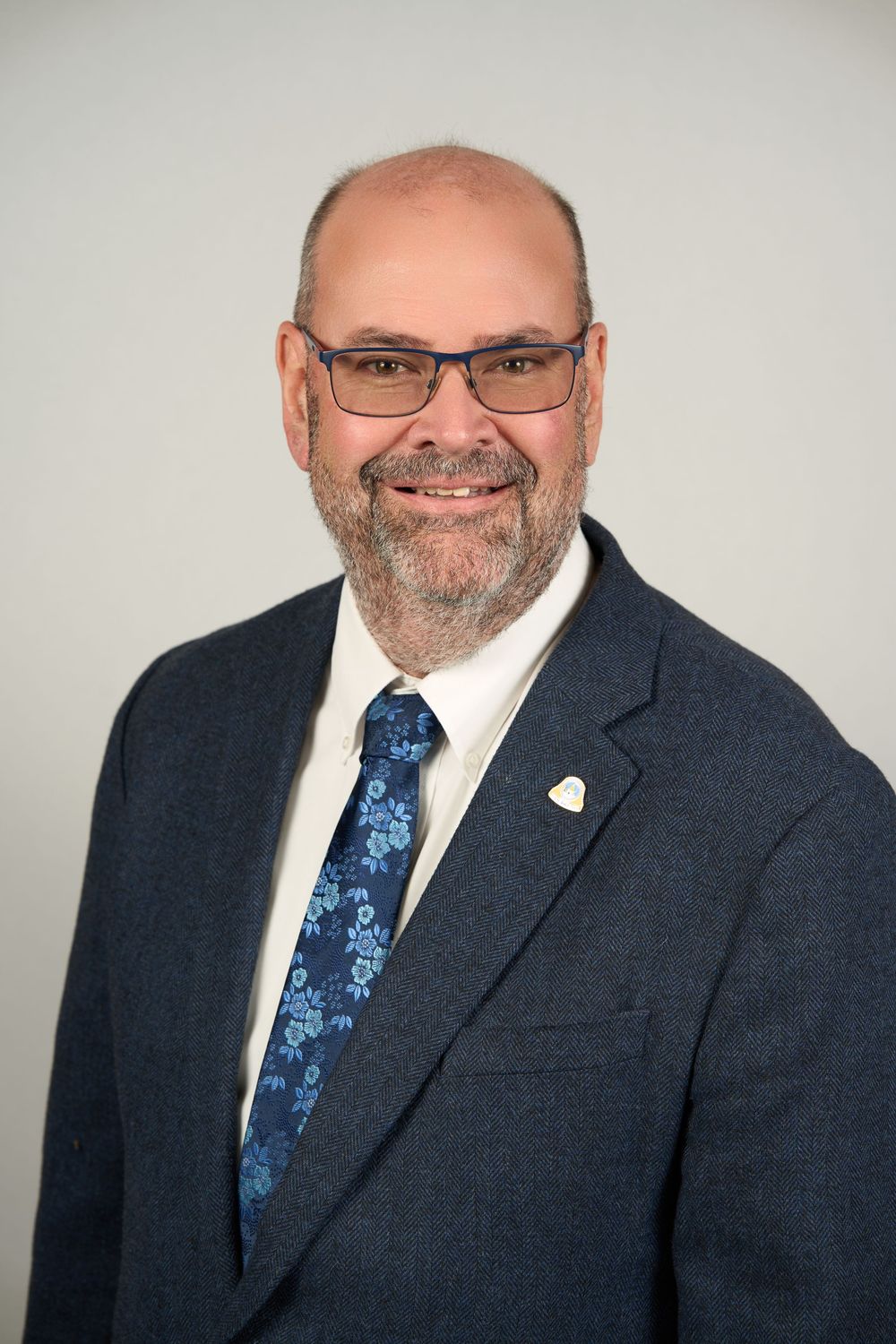 Professional headshot of a smiling bald man with a beard, wearing glasses, a navy suit, and a floral blue tie.