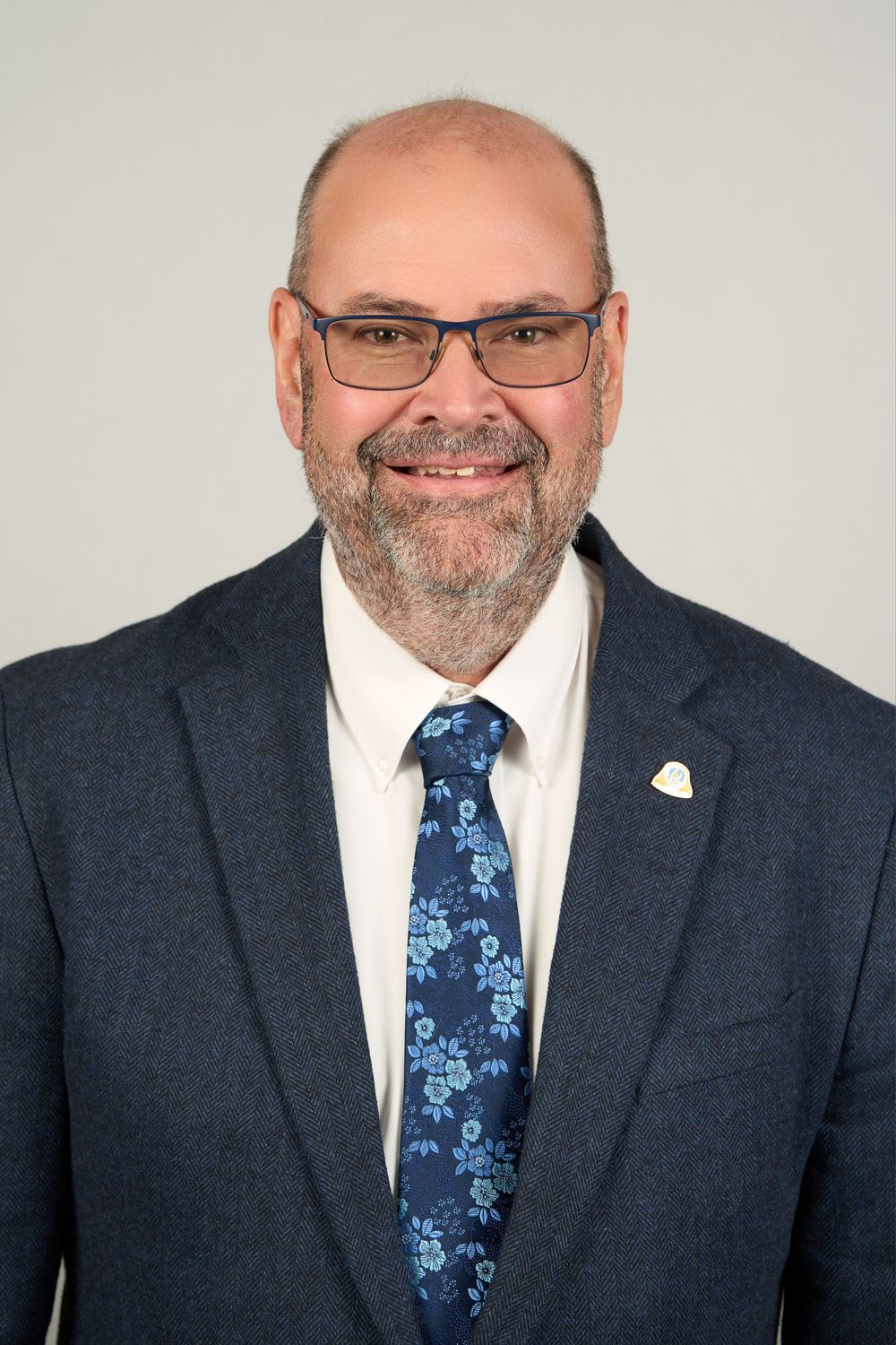 Professional headshot of a smiling bald man with glasses and beard wearing a navy suit and floral tie.